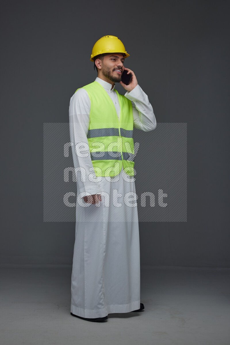 A Saudi man wearing Thobe with a yellow safety vest and white helmet standing and talking in the phone eye level on a gray background