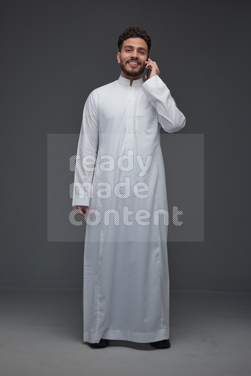A Saudi man wearing Thobe and talking in the phone while standing and making different poses eye level on a gray background