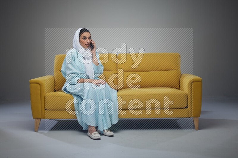 A Saudi woman wearing a light blue Abaya and a white head scarf sitting on a yellow sofa and talking in the phone while making different poses eye level on a grey background