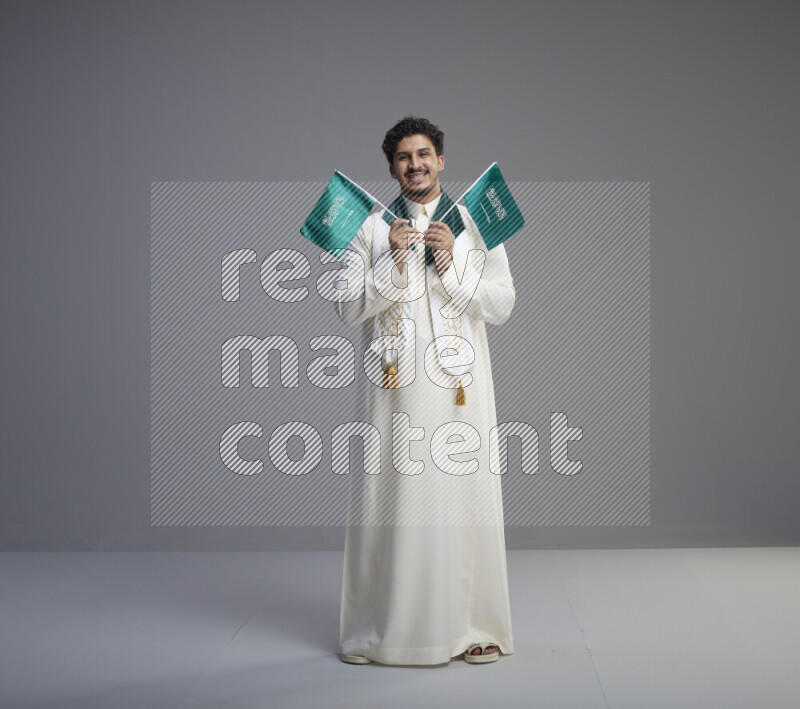 A Saudi man standing wearing thob and saudi flag scarf and holding small saudi flag on gray background