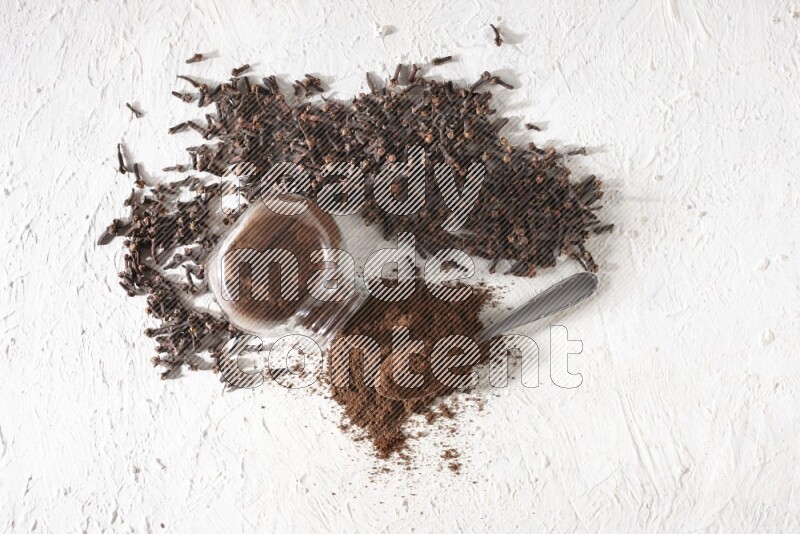 A flipped glass spice jar and a metal spoon full of cloves powder and powder came out of the jar with cloves spread on textured white flooring
