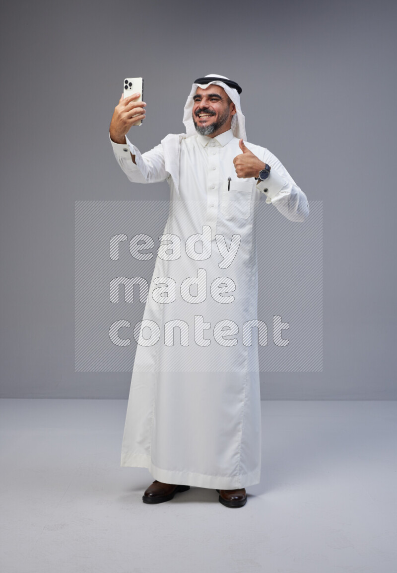 Saudi man Wearing Thob and white Shomag standing taking selfie on Gray background