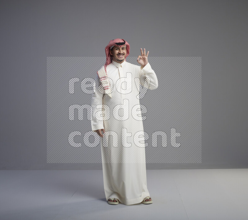 A Saudi man standing wearing thob and red shomag interacting with the camera on gray background