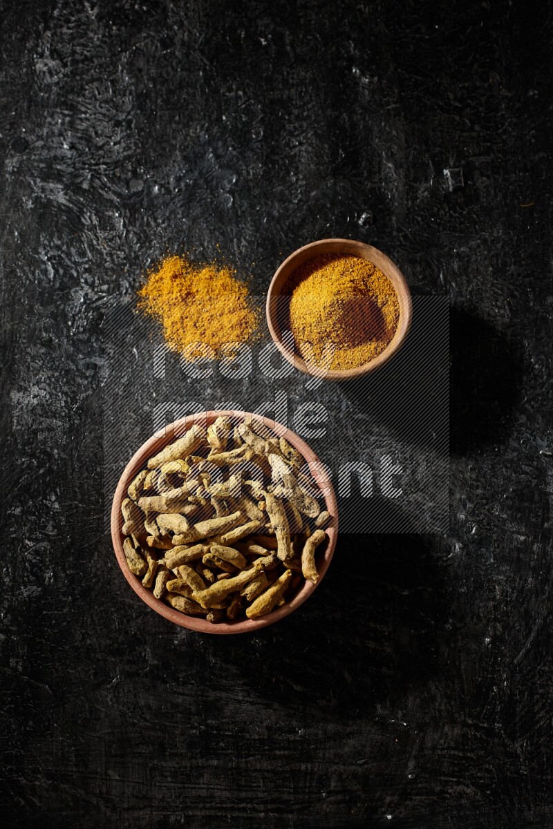 2 wooden bowls, one full of turmeric powder and the other full of dried turmeric whole fingers on a textured black flooring