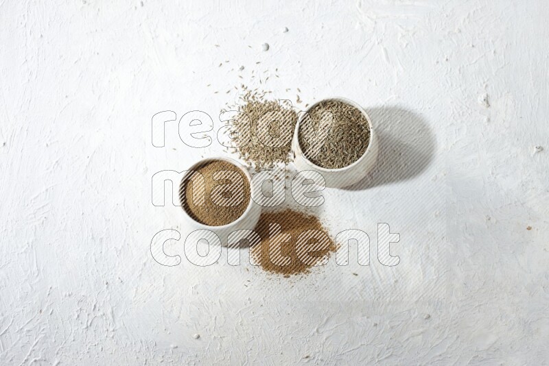 2 beige bowls full of cumin seeds and powder with spilled powder and seeds on textured white flooring