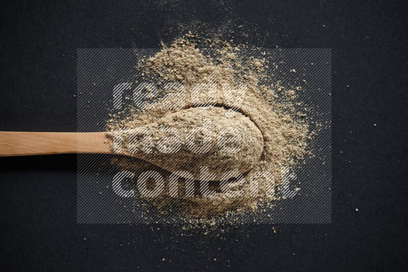 A wooden spoon full of cardamom powder and powder spreaded on black flooring