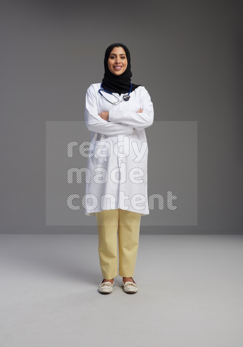 Saudi woman wearing lab coat with stethoscope standing with crossed arms on Gray background