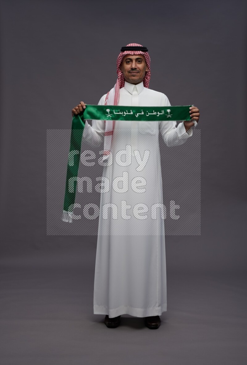Saudi man wearing thob and shomag standing holding Saudi flag on gray background
