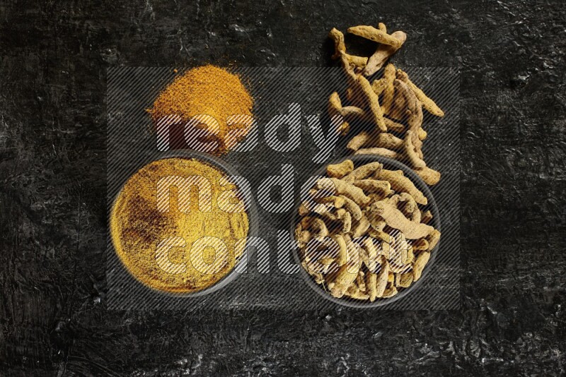 2 pottery black bowls, one full of turmeric powder and the other full of dried turmeric whole fingers on a textured black flooring