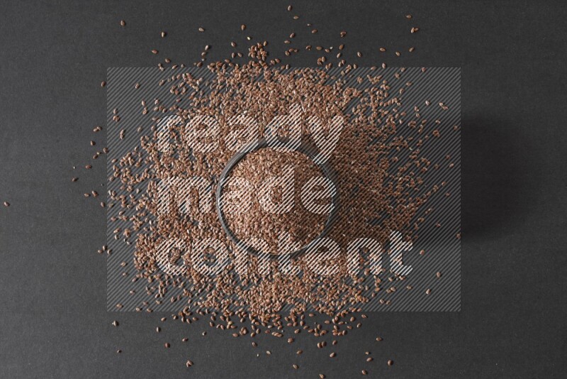 A black pottery bowl full of flaxseeds surrounded by the seeds on a black flooring