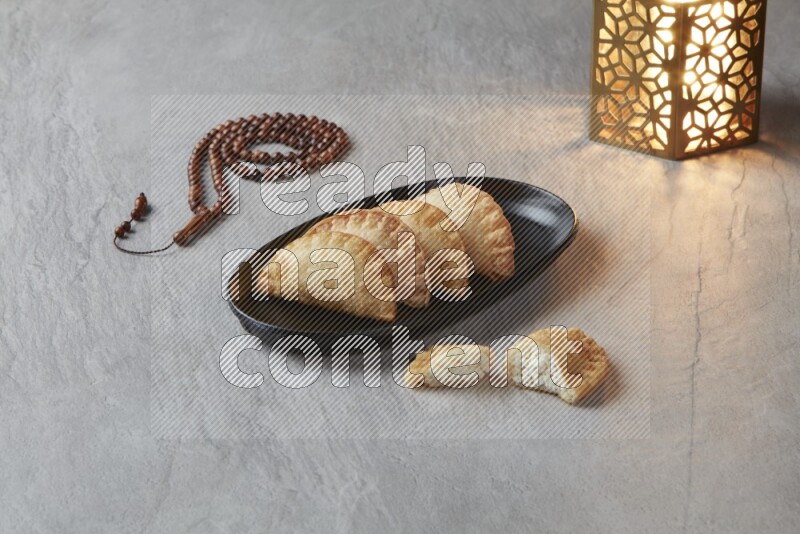 Four fried sambosas in an oval shaped black plate, beside a cut cheese sambosa, a brown misbaha and a golden lantern on a gray background