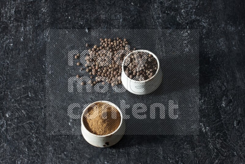2 beige bowls, one full of allspice powder and the other full of whole balls and both spread on a textured black flooring