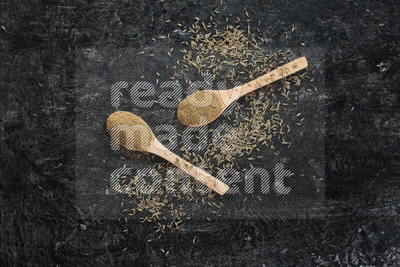 2 wooden spoons full of cumin powder with spreaded seeds on textured black flooring