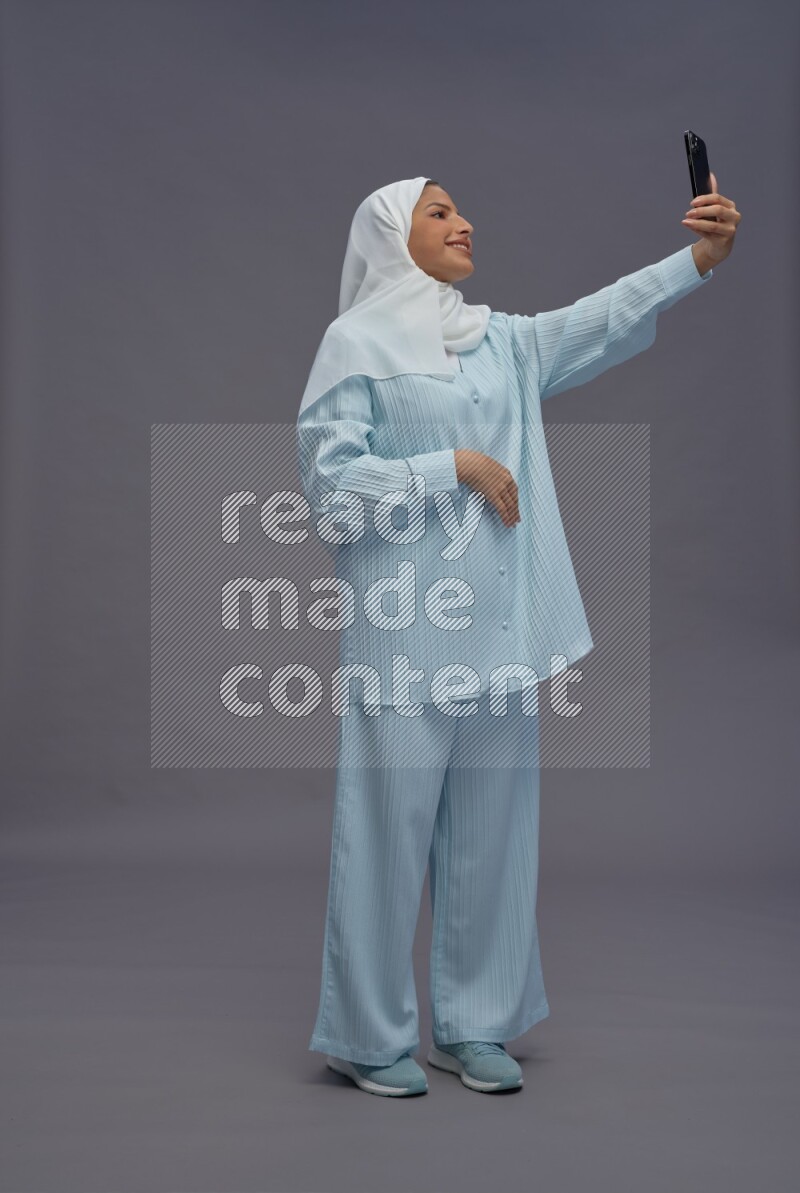 Saudi woman wearing hijab clothes standing taking selfie on gray background