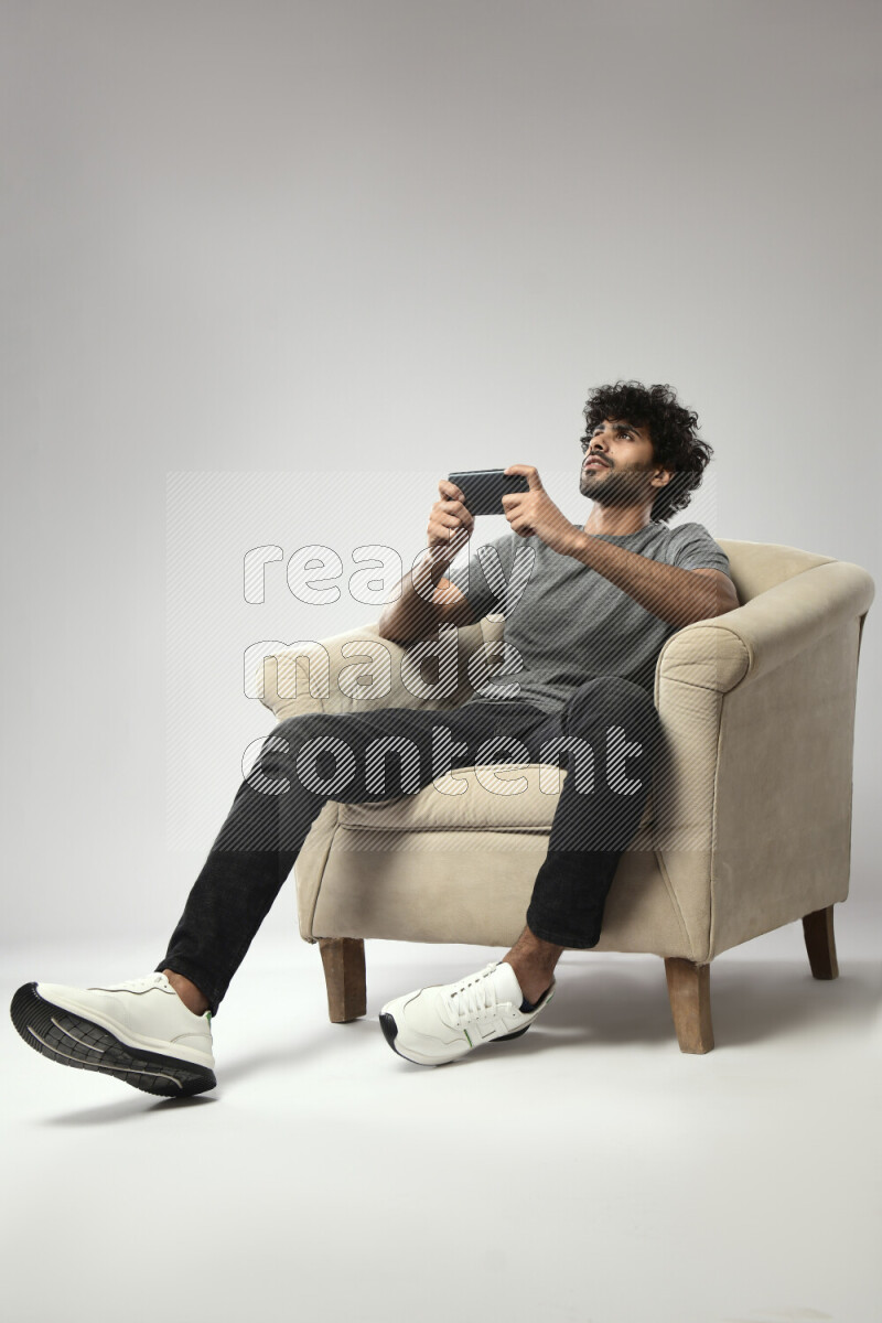 A man wearing casual sitting on a chair gaming on the phone on white background