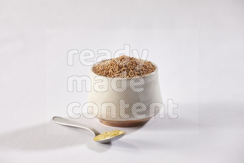 A beige pottery bowl full of mustard seeds and a metal spoon full of mustard paste on white flooring