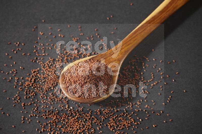 A wooden ladle full of garden cress seeds and seeds spread beside it on a black flooring