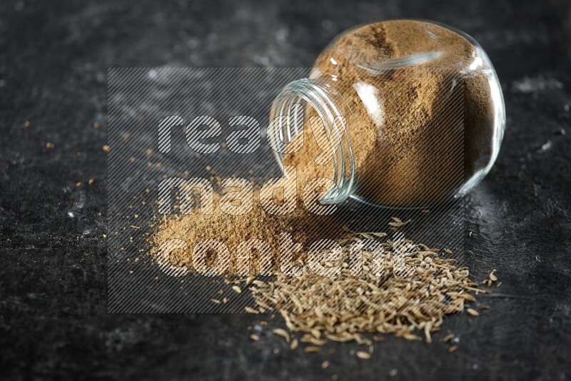 A flipped glass spice jar full of cumin powder with spilled powder and cumin seeds on a textured black flooring