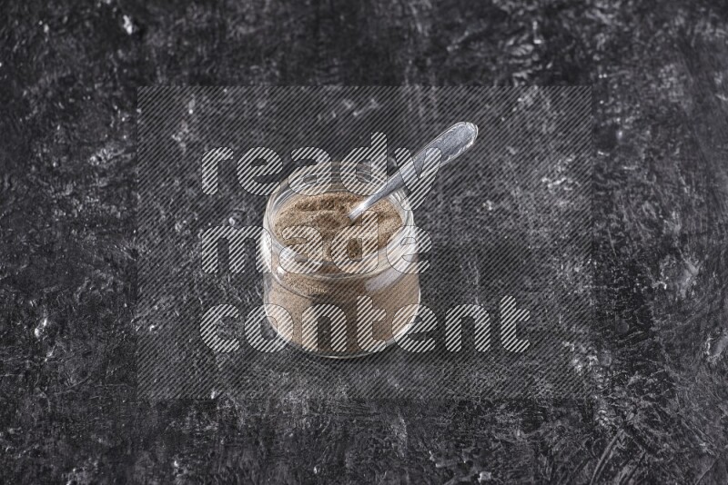 A glass jar full of black pepper powder and a metal spoon on a textured black flooring