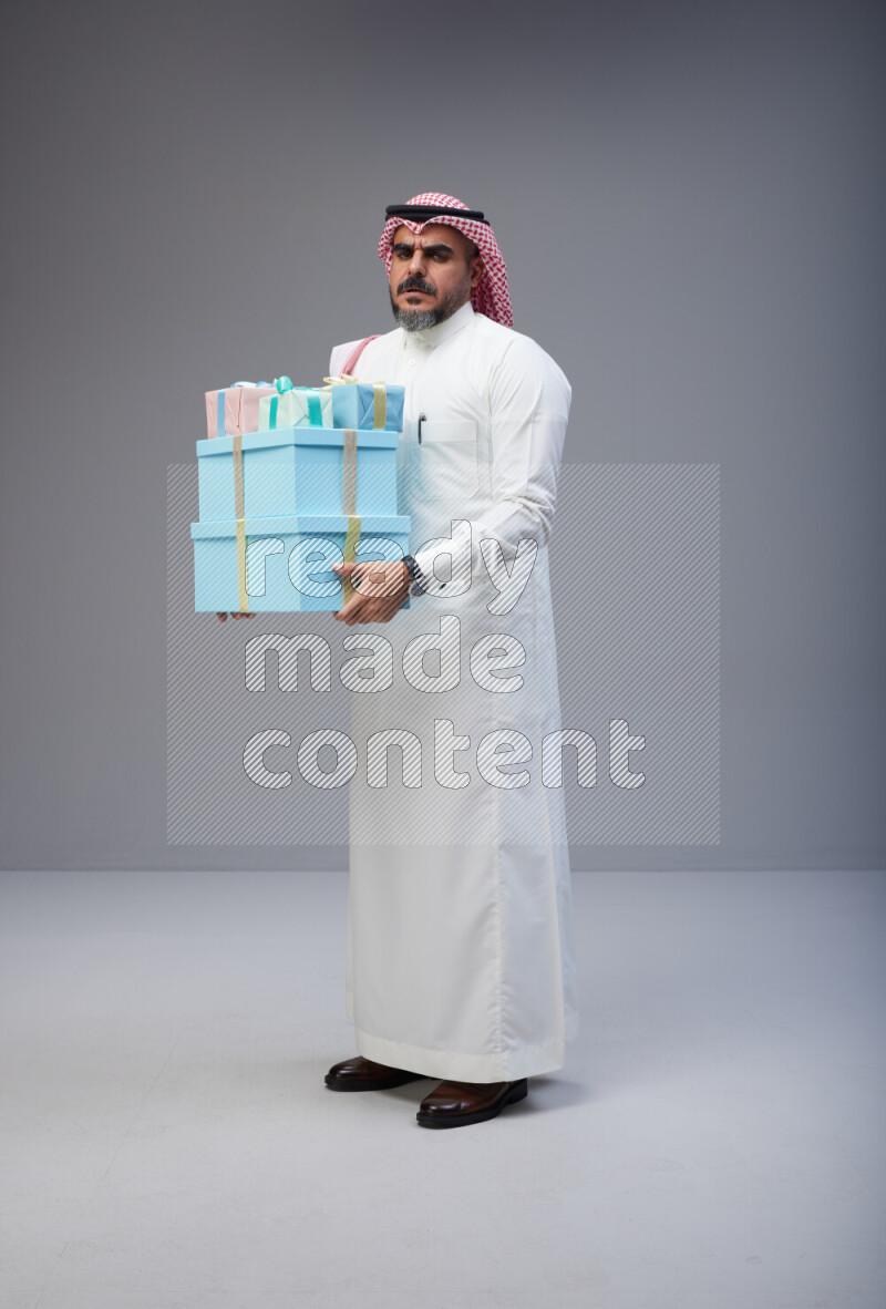 Saudi man Wearing Thob and red Shomag standing holding gift box on Gray background