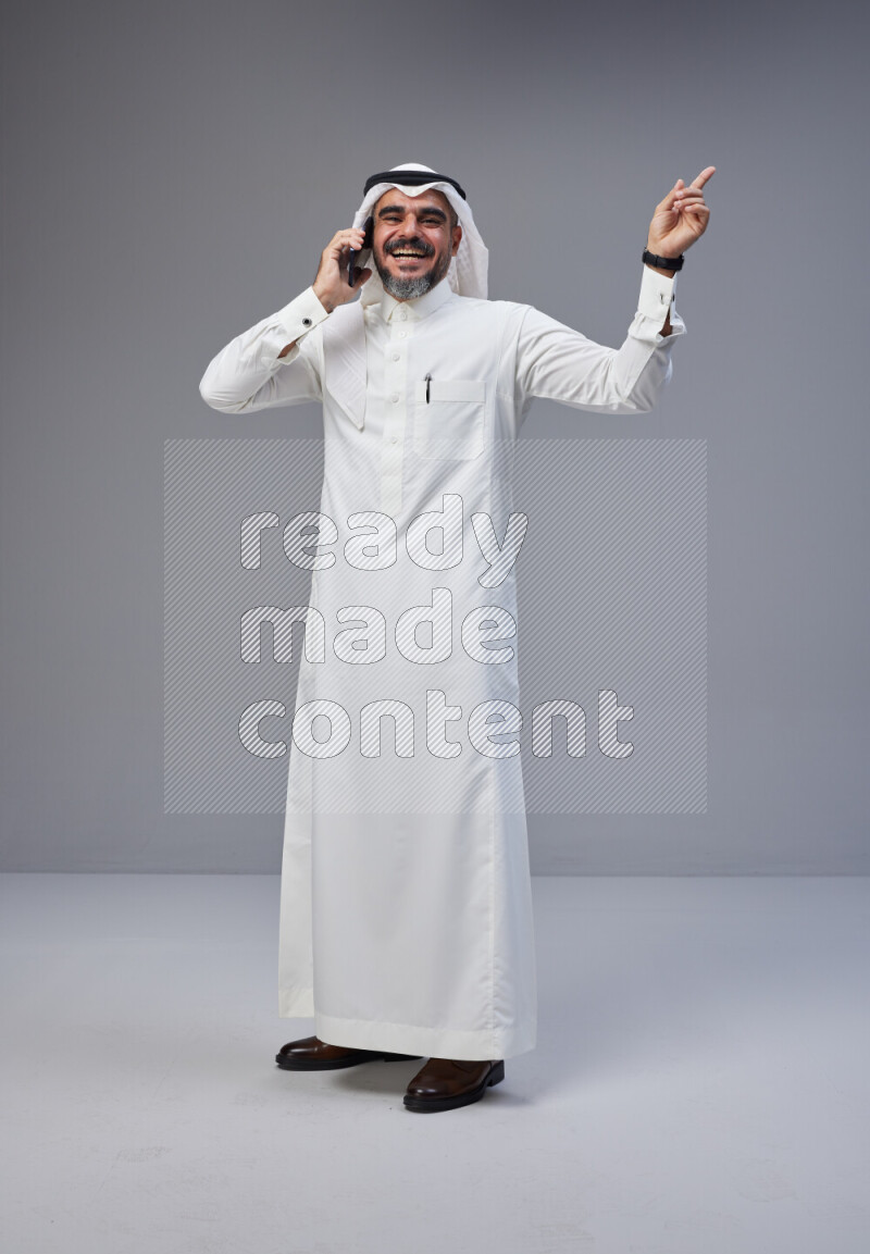 Saudi man Wearing Thob and white Shomag standing talking on phone on Gray background