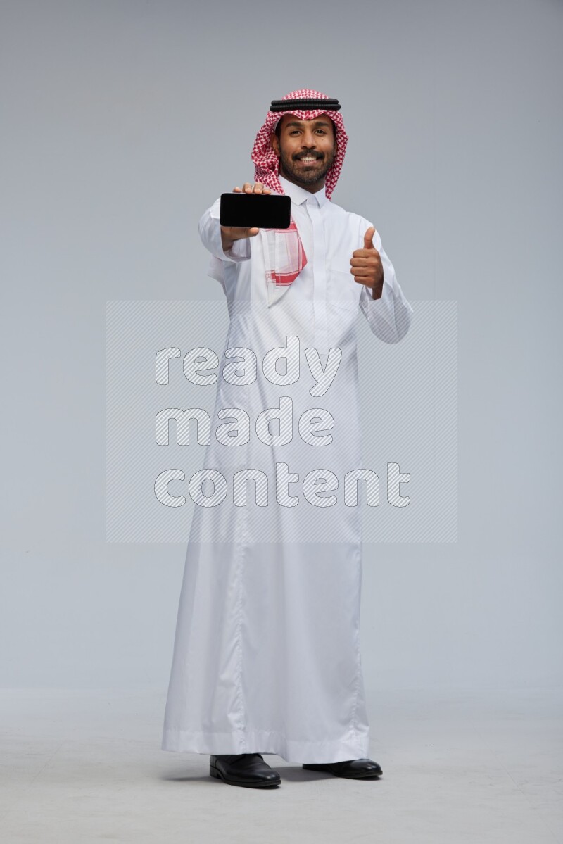 Saudi man Wearing Thob and shomag standing showing phone to camera on Gray background