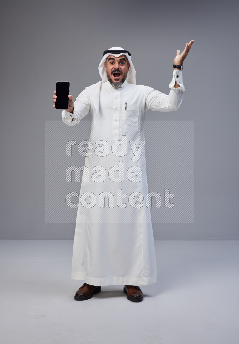 Saudi man Wearing Thob and white Shomag standing showing phone to camera on Gray background