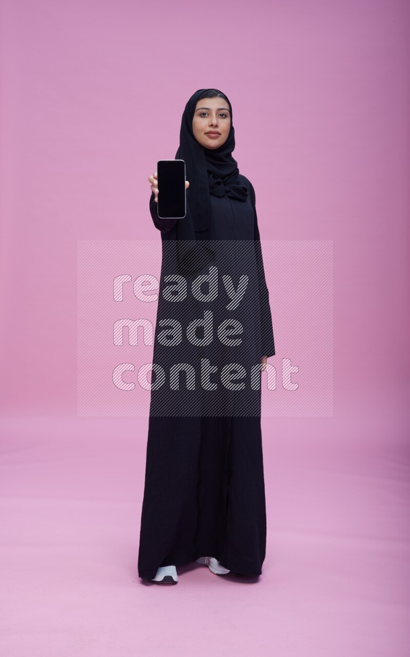 Saudi woman wearing Abaya standing showing phone to camera on pink background