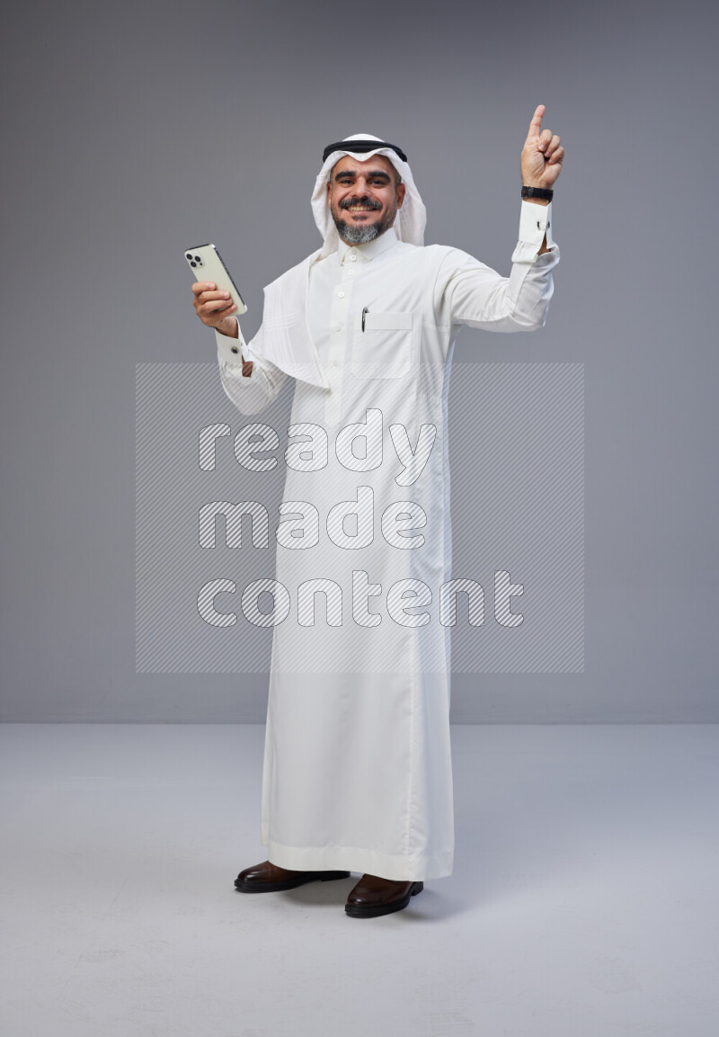 Saudi man Wearing Thob and white Shomag standing texting on phone on Gray background