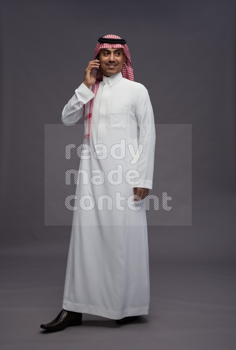 Saudi man wearing thob and shomag standing talking on phone on gray background