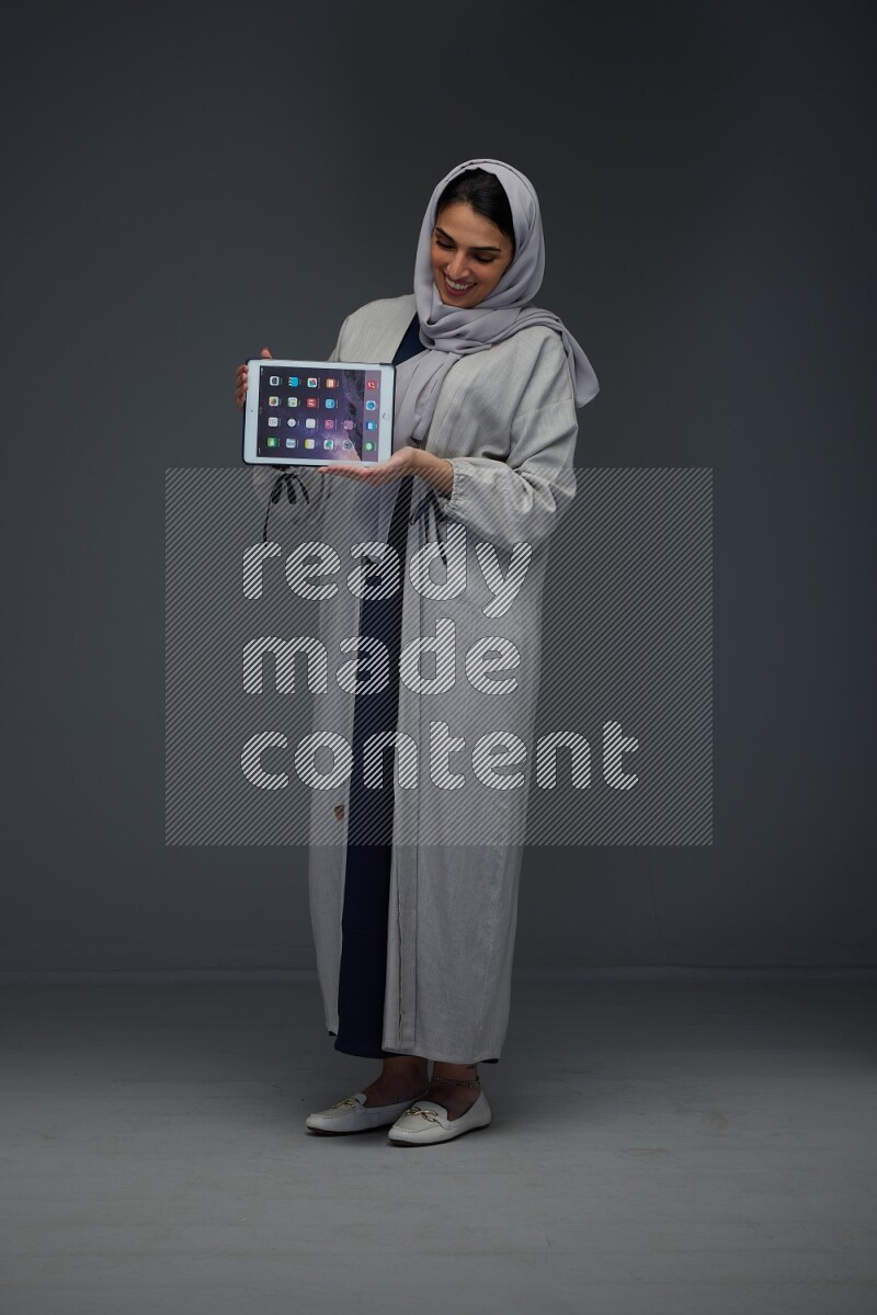 A Saudi woman wearing a light gray Abaya and head scarf standing and showing the phone's screen eye level on a grey background