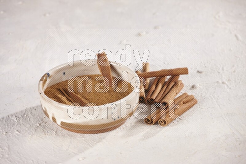 Ceramic bowl full of cinnamon powder with cinnamon sticks on the side on white background