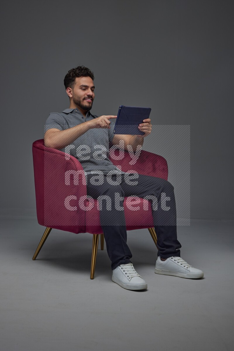 A man wearing casual and using his tablet while sitting on a burgundy chair eye level on a gray background