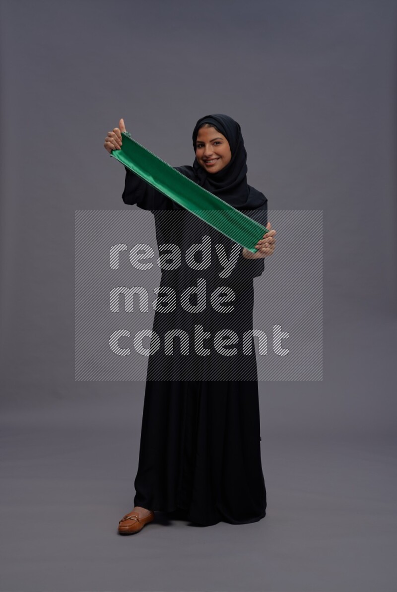 Saudi woman wearing Abaya standing holding Saudi flag on gray background