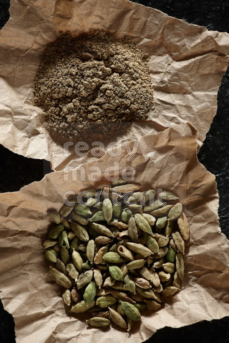 Cardamom seeds and cardamom powder in 2 crumpled pieces of paper on textured black flooring