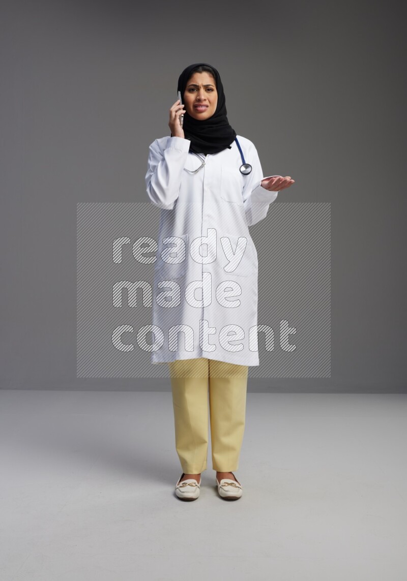 Saudi woman wearing lab coat with stethoscope standing talking on phone on Gray background