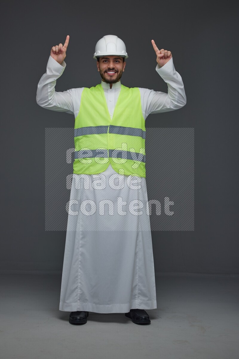 A Saudi man wearing Thobe with a yellow safety vest and white helmet standing and pointing different angles eye level on a gray background