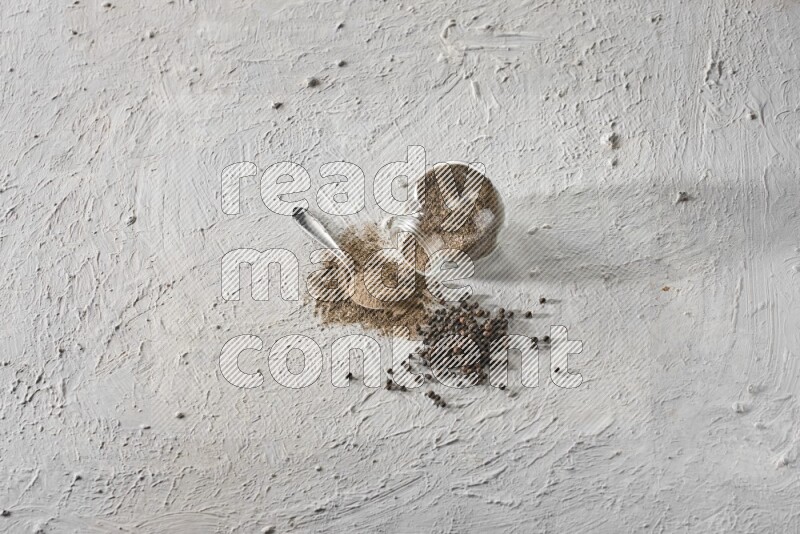 Flipped glass spice jar full of black pepper powder with a metal spoon and black pepper beads spread on a textured white flooring