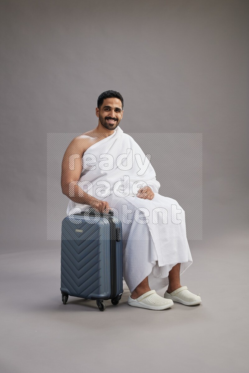 A man wearing Ehram sitting on chair holding traveling bag on gray background
