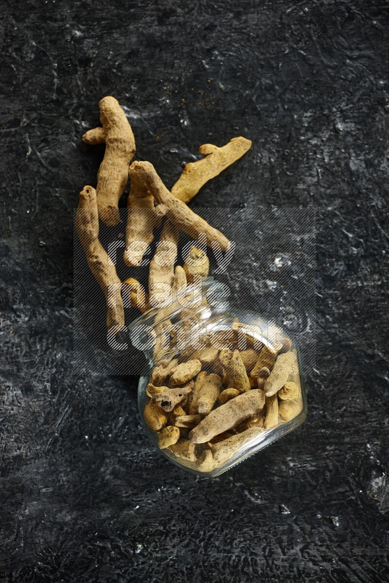 A flipped glass spice jar full of dried turmeric whole fingers and fingers fell out of it on textured black flooring