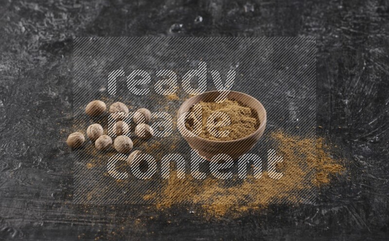 A wooden bowl full of nutmeg powder with whole seeds and sprinkled powder beside it on a textured black flooring
