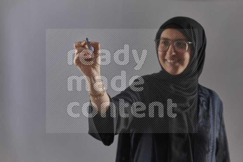 Woman in black abaya, hijab and glasses holding a marker pen to write on a transparent board with different reactions and expressions on grey background