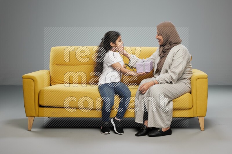 A girl sitting on a yellow sofa and giving a gift to her mother on gray background