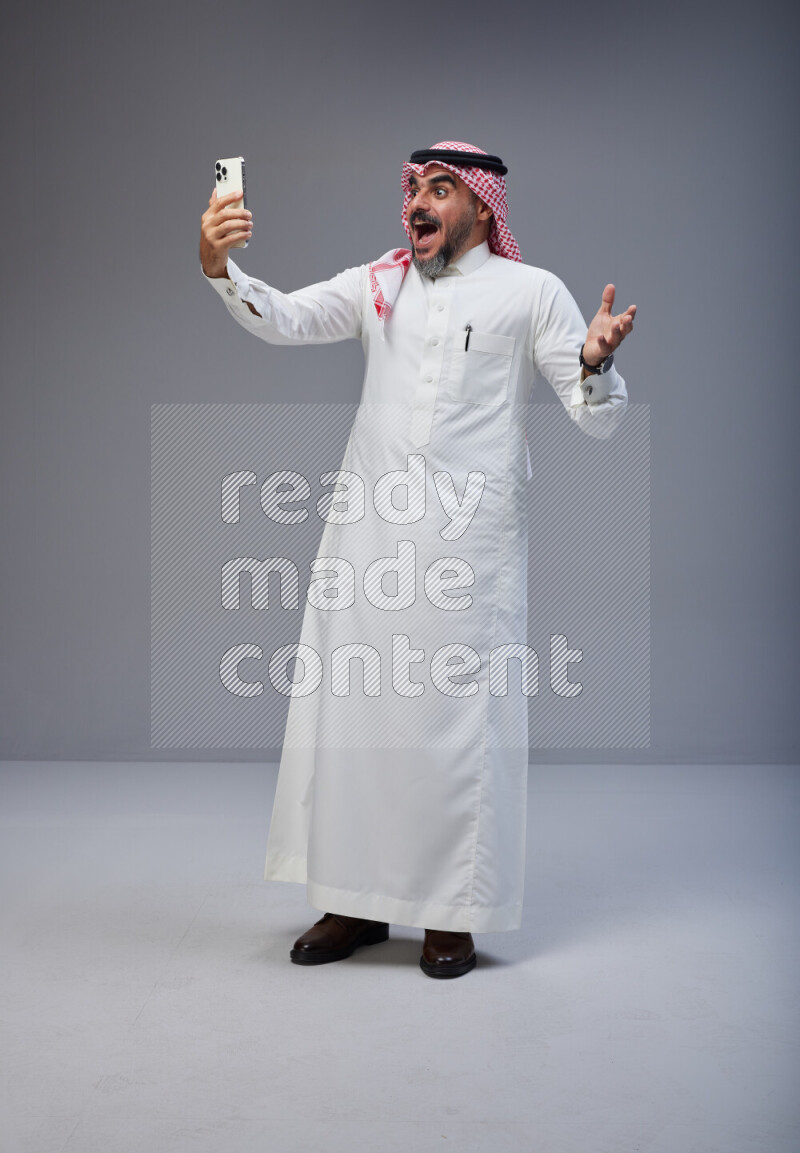 Saudi man Wearing Thob and red Shomag standing taking selfie on Gray background
