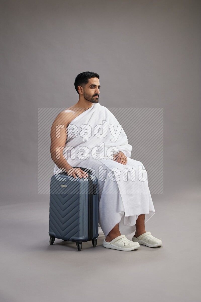 A man wearing Ehram sitting on chair holding traveling bag on gray background