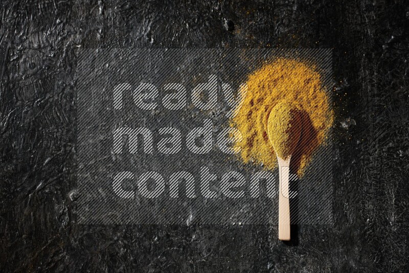 A wooden spoon full of turmeric powder on textured black background