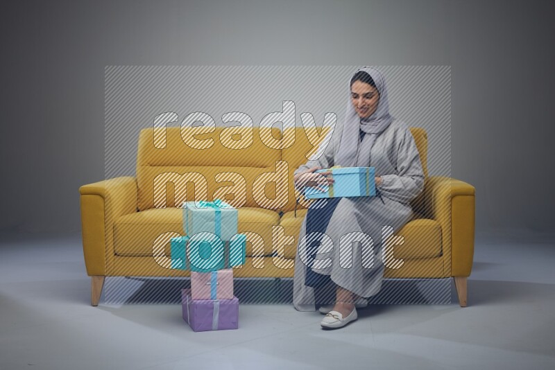 A Saudi woman wearing a light gray Abaya and head scarf sitting on a yellow sofa holding a gift box beside multi color gift boxes eye level on a grey background