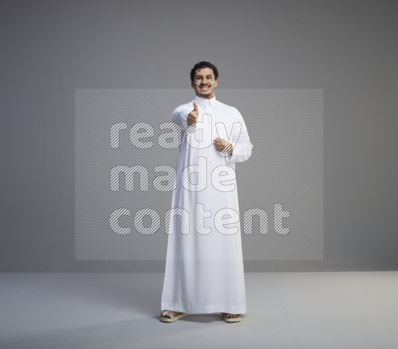 A Saudi man standing wearing thob with face painting interacting with the camera on gray background