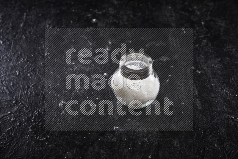 A glass jar full of coarse sea salt crystals on black background
