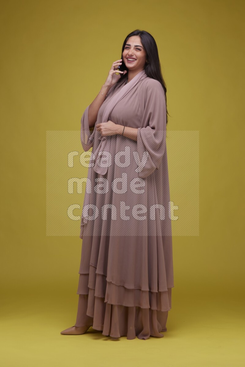 A woman Calling on a Yellow Background wearing Brown Abaya